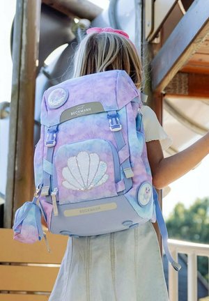 Bambino con zaino pastello viola e blu con design di conchiglie marine e tasche laterali, in piedi vicino a una struttura da gioco in legno all'aperto.