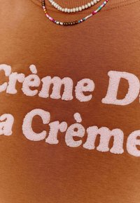 Hnědá mikina s měkkým povrchem, na které je vyšitý růžový text "Crème De la Crème." Doplněk zahrnuje náhrdelník z vícebarevných korálků.
