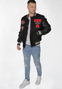 Svart varsityjacka med läderärmar, röda märken, vita ränder på ärmsluten, bärs över en vit t-shirt, i kombination med ljusblå slitna jeans.