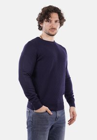 Maglione blu navy con texture, scollo a giro e maniche lunghe. Il modello lo indossa con jeans blu, evidenziando un design aderente e un motivo sottile.