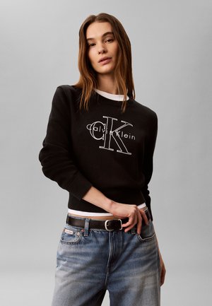 Młoda kobieta ubrana w czarny sweter Calvin Klein i wyblakłe niebieskie jeansy, pozująca z jedną ręką blisko paska na szarym tle.