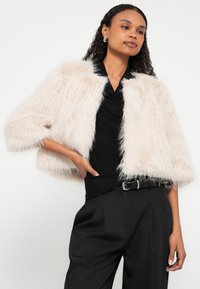 Svetlo roza umetno krzno jakna s kratkimi rokavi in ovratnikom, nošena čez črno majico, kombinirana s črnimi plisiranimi hlačami in pasom.