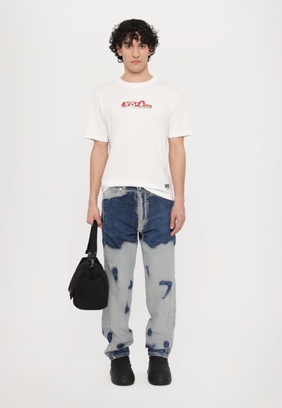 T-shirt blanc en coton avec le logo "Evisu" rouge, jeans en denim délavé bleu et gris, sac noir et chaussures noires, avec une coupe décontractée.