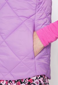 Gilet matelassé violet clair avec un motif de couture diagonal, doté d'une poche latérale ; associé à une chemise à manches longues rose et une jupe à fleurs.