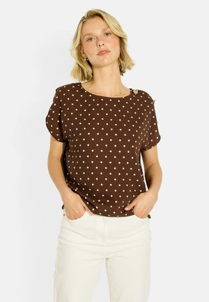 Mujer de pelo rubio corto lleva una blusa marrón de lunares y pantalones blancos, de pie con las manos en los bolsillos sobre un fondo liso y claro.