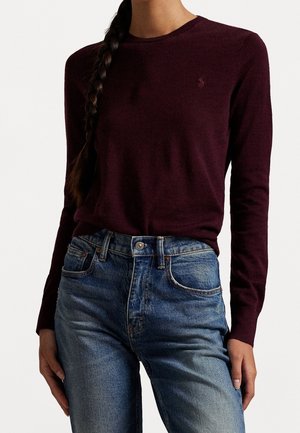 Person iført en mørk bordeaux langærmet sweater og blå højtaljede jeans med en flettet ponyhale over den ene skulder.