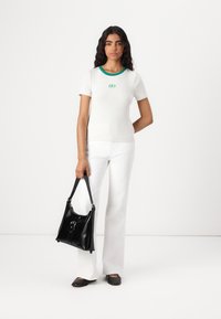 Camisa gris de manga corta con cuello verde y logo, combinada con pantalones acampanados blancos. Bolso negro con herrajes de acento, llevado en la mano izquierda.