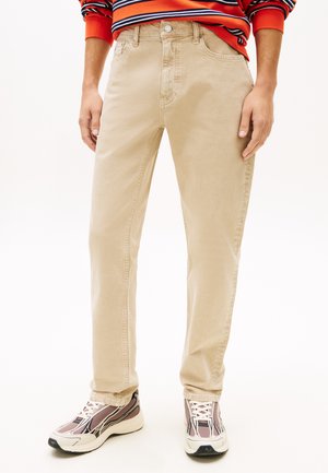 Persona vistiendo pantalones beige de pierna recta, suéter de rayas naranjas y zapatillas de colores claros, de pie con las manos relajadas a los lados.