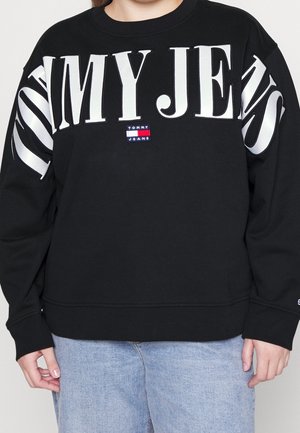 Sort sweatshirt med stor hvid tekst "TOMMY JEANS" hen over brystet og ærmerne, lille Tommy Jeans logo patch i midten, båret med blå jeans.