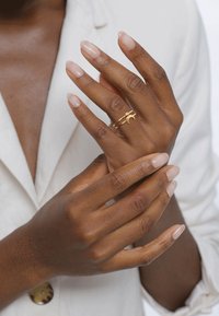 Bague en or avec un détail en croissant de lune et une petite pierre précieuse, portée sur une main avec des ongles nus et brillants, sur un fond doux et clair.