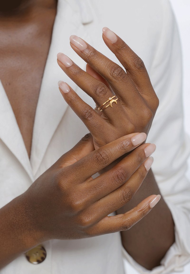 Bague en or avec un détail en croissant de lune et une petite pierre précieuse, portée sur une main avec des ongles nus et brillants, sur un fond doux et clair.