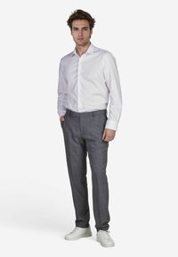 Ein junger Mann steht selbstbewusst da, trägt ein weißes Hemd und graue gemusterte Hosen, kombiniert mit weißen Sneakers, vor einem schlichten Hintergrund.
