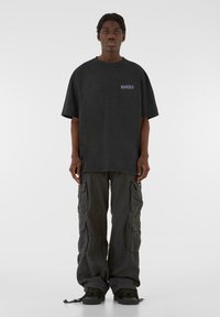 Svart oversized t-skjorte i bomull med lilla grafikk, parret med mørke cargo-bukser med flere lommer og løs passform. Svart joggesko.