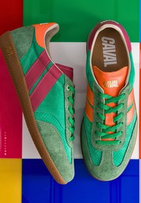 Paire de baskets en daim vert avec des détails orange et rose, des lacets verts et des semelles en caoutchouc marron sur une surface carrelée colorée.