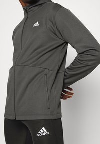 Chaqueta deportiva gris con cremallera y capucha, fabricada con un tejido texturizado, presentada con bolsillos laterales y el logo blanco de Adidas en el pecho.