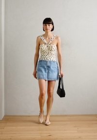 White top with black polka dots, twisted design, paired with a light blue denim skirt. Carrying a small black handbag. Light wooden floor.
