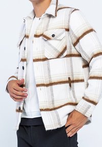 Rutig skjorta i vita och bruna nyanser, gjord av mjukt tyg. Har två bröstfickor, svarta knappar och en krage.