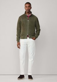 Chaqueta de ante verde oliva ajustada, pantalones blancos y zapatos oscuros de vestir sin cordones. La chaqueta tiene un cuello alto y cierre de cremallera.