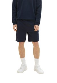 Muž v námořnických modrých cargo šortkách, odpovídajícím dlouhém rukávu, bílých ponožkách a bílých teniskách, stojící na bílém pozadí.