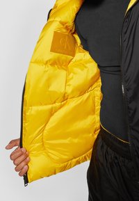 Svart ytterjacka med gul satininteriör. Har vadderade sektioner och en Calvin Klein-logotyp på lappen. Dragkedjan är stängd på vänster sida.