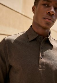 Tricou polo maro dintr-un material texturat, cu guler și trei butoane, surprins dintr-un unghi apropiat care evidențiază detaliile tricoului.