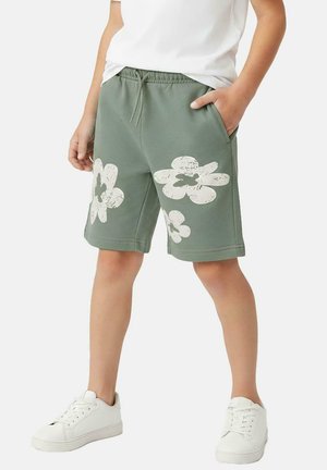 Groene katoenen korte broek met grote witte bloemenprints op beide pijpen, voorzien van een elastische tailleband en zijzakken, gecombineerd met witte sneakers.