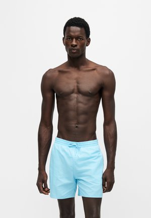 Uomo muscoloso dalla pelle scura in piedi di fronte, indossa pantaloncini da bagno azzurri su uno sfondo bianco semplice.