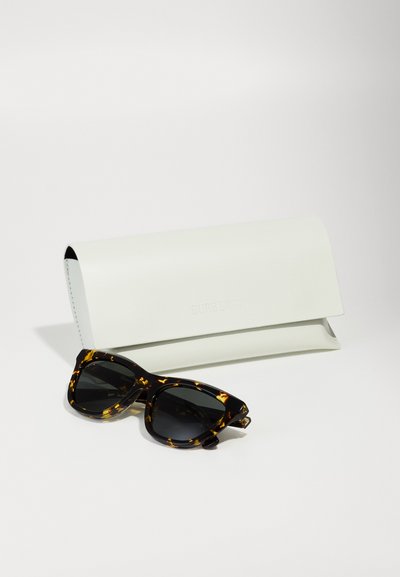 Sunkiai su akinių rėmeliais ir tamsiais lęšiais, turinčiais drąsią kampuotą formą, šalia lygios baltos odinės dėklo su įspaustu žodžiu "Burberry."