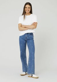 Jean en denim bleu clair évasé avec des motifs de vache brodés en blanc, associé à un t-shirt blanc uni et des baskets blanches avec des rayures noires.