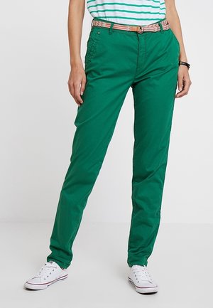 Personne portant un pantalon vert, une chemise rayée, une ceinture à motif et des baskets blanches, se tenant devant un fond clair uni.