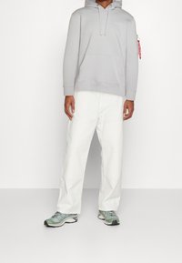 Sudadera gris con bolsillo frontal y capucha ajustable, combinada con pantalones blancos holgados y zapatillas deportivas verdes.