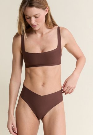 Donna che sistema un costume da bagno marrone a due pezzi con scollo quadrato e slip a vita alta, in piedi contro uno sfondo chiaro uniforme.