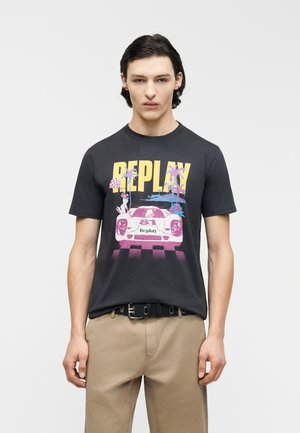 Νεαρός άνδρας που φοράει μαύρο μπλουζάκι με γραφικό σχέδιο vintage αυτοκινήτου, φοινικόδεντρα και την επιγραφή "REPLAY", συνδυασμένο με μπεζ παντελόνι και μαύρη ζώνη.