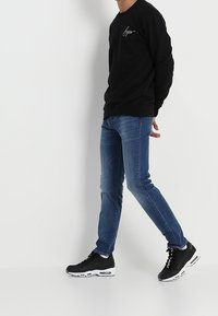 Czarny dres z logo w stylu pisma, niebieskie dopasowane dżinsy i czarne trampki z białymi akcentami, na jednolitym tle.