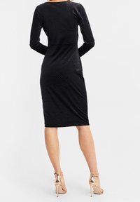 Musta vartalonmyötäinen mekko, jossa pitkät hihat ja geometrinen tekstuurikuvio. Yhdistetty metallinsävyisiin korkokenkiin.