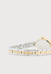 Guld- og sølvmetal kæde til håndleddet med synlig Timex ur bagbeklædning, der hviler på en hvid overflade.