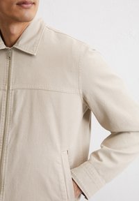 Beige lightweight jacket with a collar, front zip closure, and side pockets. Features a smooth texture and minimalistic design.