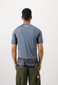 Camiseta deportiva gris de manga corta, con una sección inferior negra con patrón y un logo en la parte superior de la espalda. Textura suave.