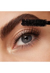 Brosse de mascara noir peignant des cils recourbés, révélant un gros plan d'une iris bleu-gris contre un teint de peau pêche illuminé.