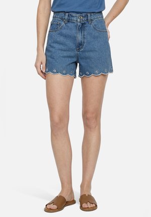 Frau trägt blaue Denim-Shorts mit wellenförmigem Saum und Lochstickerei, kombiniert mit braunen Slip-On-Sandalen und einem blauen Oberteil.