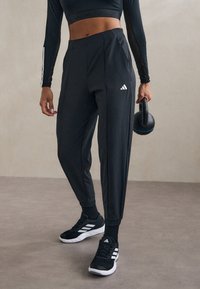 Zwarte sportieve joggings met aansluitende enkels, voorzien van subtiele plooien, zijzakken en een wit logo, gecombineerd met zwarte sportschoenen.