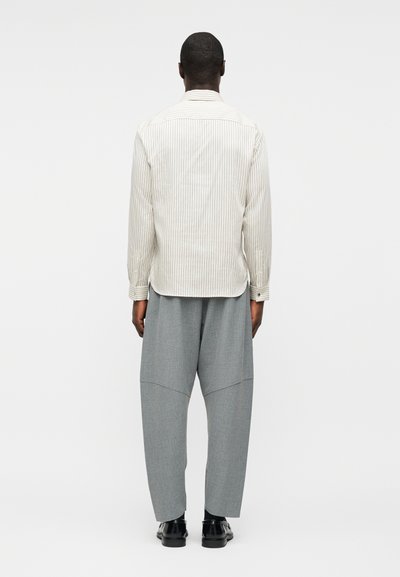 Άνδρας που κοιτάζει προς τα πίσω, φοράει λευκό ριγέ πουκάμισο με μακρυά μανίκια, φαρδιά γκρι παντελόνια με ραφή στο γόνατο και μαύρα επίσημα παπούτσια.