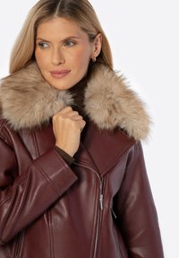 Veste en cuir bordeaux avec un col en fausse fourrure beige duveteux, dotée d'une fermeture éclair et de coutures angulaires. Texture lisse avec un design structuré.