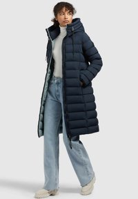 Navy puffermantel med huva, vertikal quiltad design, framdragkedja och justerbara snoddar, i kombination med ljusblå jeans och krämfärgade stövlar.