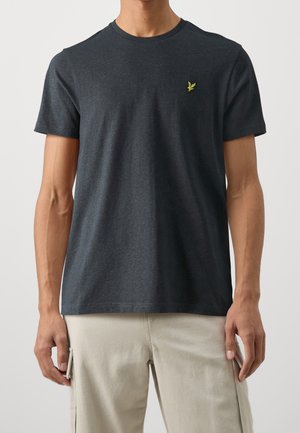 Mann trägt dunkelgraues kurzärmeliges T-Shirt mit kleinem gelbem Vogel-Logo auf der Brust und hellbeige Cargohosen, gezeigt von Hals bis Hüfte.