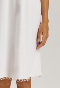 Vestido de algodón blanco con un dobladillo de encaje en forma de concha y una silueta ligeramente acampanada. La mano descansa sobre la tela, mostrando su textura suave.