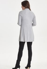 Cardigan grigio con texture a coste e retro svasato, dotato di colletto. Abbinato a pantaloni neri aderenti e stivali neri con tacco.