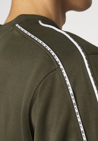 Olive green t-shirt with a textured fabric, featuring white striped accents along the shoulder seams and logo detailing.