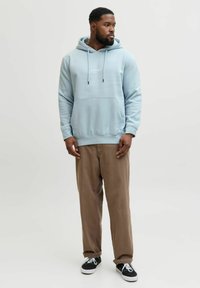 Sweat à capuche bleu clair avec une poche avant et un cordon de serrage, associé à un pantalon marron et des baskets noires. Le sweat à capuche présente un logo blanc sur la poitrine.