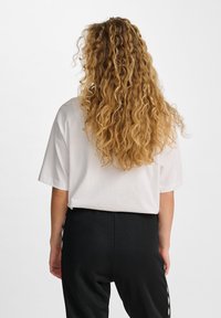 T-shirt bianca a maniche corte, con una texture liscia, abbinata a pantaloni neri con dettagli laterali. Capelli ricci di lunghezza media visibili.